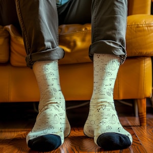 May include: Off-white socks with black heels and toes, featuring a repeating pattern of black mathematical equations and diagrams. The socks are worn by a person sitting on a yellow sofa.