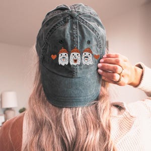 May include: A faded teal baseball cap with an embroidered design of three ghost figures wearing orange beanies and holding coffee mugs. The design includes orange hearts on either side of the ghosts. The cap is worn by a person with blonde hair.