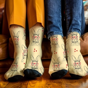 May include: A pair of cream-colored socks with a black and white bunny wearing red glasses pattern. The socks are worn by two people sitting on a brown couch.