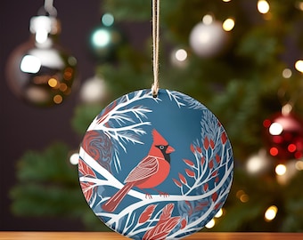 May include: A round ceramic ornament with a blue background and a white silhouette of a cardinal bird perched on a branch. The bird is red with a black beak and eye. The ornament is decorated with white branches and red berries.