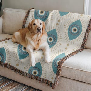 May include: A golden retriever dog sits on a cream-colored throw blanket with a blue and gold eye pattern. The blanket has fringed edges.