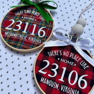 May include: Two round ornaments with wooden embroidery hoops. One has a red, green, and black plaid pattern with the text "THERE'S NO PLACE LIKE HOME" and "23116 MECHANICSVILLE, VIRGINIA". The other has a red and black plaid pattern with the text "23106 MANQUIN, VIRGINIA".