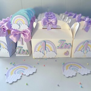May include: Rainbow-themed party favor boxes and cones with a purple ribbon and the name "Zoe" on them. The boxes are white with a rainbow, clouds, and the number "1" on them. The cones are white with a rainbow and the name "Zoe" on them.
