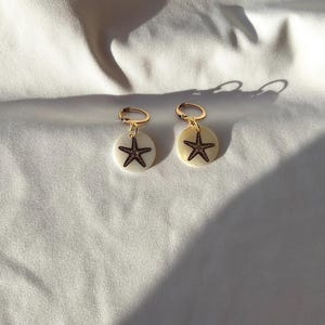 Op de afbeelding: Een paar gouden creolen met een witte schelpbedel met een zwart zeesterontwerp.