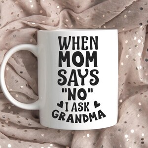 Puede incluir: Taza de cerámica blanca con asa curva. La taza presenta texto negro que dice "WHEN MOM SAYS 'NO' I ASK GRANDMA" con acentos de corazón. La taza es un regalo para una abuela.