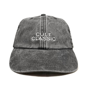 May include: A grey baseball cap featuring the embroidered text "CULT CLASSIC" in white. The cap has a curved peak and a button on top. The hat is made of a faded, textured material.