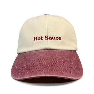 May include: A baseball cap featuring a cream-colored crown and a distressed maroon brim. The text "Hot Sauce" is embroidered in dark red thread across the front center of the crown. The cap has a structured six-panel design with visible stitching.
