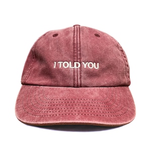 May include: A maroon baseball cap with a washed, vintage-style finish. The front panel features the text "I TOLD YOU" embroidered in white thread. The cap has a curved brim with visible stitching and a structured crown with eyelet ventilation holes.