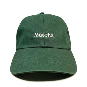 May include: A dark forest green baseball cap featuring the word Matcha embroidered in white thread across the front centre. The cap has a structured six-panel crown, a curved brim with visible stitching, and a button at the top centre.
