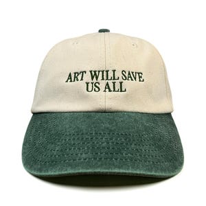 Peut inclure: Une casquette de baseball avec une calotte crème et une visière vert foncé. L'avant de la casquette présente le texte brodé "ART WILL SAVE US ALL" en fil vert foncé. La casquette possède un panneau avant structuré et une visière incurvée.