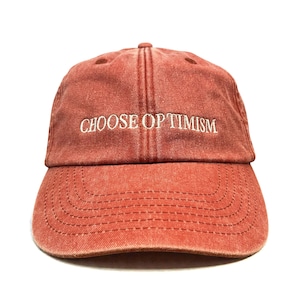 Può includere: Un cappellino da baseball color mattone con la scritta ricamata "CHOOSE OPTIMISM" in bianco. Il cappellino ha una visiera curva e un design a sei pannelli con un bottone sulla parte superiore. Il tessuto sembra essere cotone lavato.