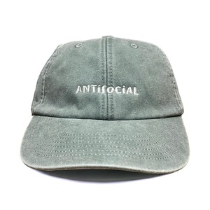 May include: A sage green baseball cap with a six-panel construction and a curved brim. The word ANTISOCIAL is embroidered in white thread across the front centre of the crown. The fabric has a washed, slightly faded appearance throughout.