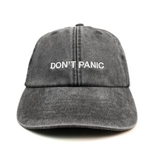 May include: A charcoal grey baseball cap with a washed, distressed finish. The front panel features the text DON'T PANIC embroidered in white capital letters. The cap has a curved brim, a six-panel construction, and a button on the top crown.