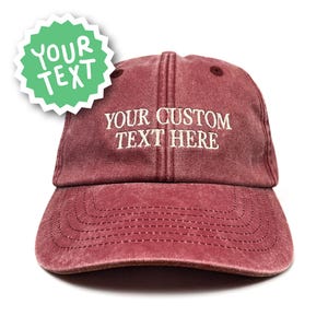 May include: A maroon baseball cap with a distressed, washed-out finish. The front features white embroidered text that reads YOUR CUSTOM TEXT HERE. A light green starburst-shaped graphic in the upper left corner displays the text YOUR TEXT.