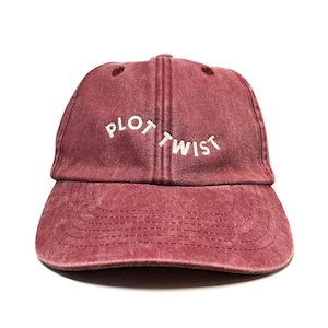 May include: A maroon baseball cap with a washed-out, vintage texture. The front panel features the text "PLOT TWIST" embroidered in white, sans-serif capital letters, arranged in a slight arch across the centre of the cap above the brim.