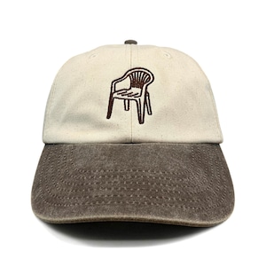May include: A baseball cap featuring a cream-coloured crown and a distressed dark brown brim. The front panel displays a brown embroidered graphic of a plastic patio chair. The cap includes a small brown button at the top centre of the crown.