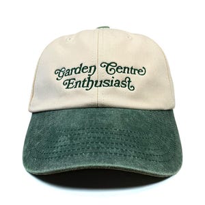 May include: A baseball cap with a cream-coloured crown and a dark green, distressed-look brim. The front of the cap features the embroidered text "Garden Centre Enthusiast" in a dark green, stylised cursive font with decorative flourishes.