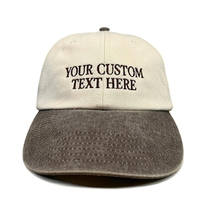 Könnte beinhalten: Eine Baseballkappe mit cremefarbener Krone und einem abgenutzten, dunkelbraunen Schirm. Auf der Vorderseite der Kappe befindet sich der gestickte Text "YOUR CUSTOM TEXT HERE" in dunkelbrauner Schrift, zentriert über die zwei Vorderteile.
