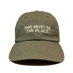 May include: An olive green baseball cap featuring the embroidered text "THIS MUST BE THE PLACE" in a light cream colour on the front panel. The cap has a curved brim with visible stitching and eyelets on the crown for ventilation and structure.
