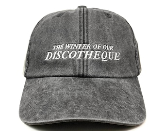May include: A charcoal grey baseball cap with a washed, distressed finish. The front features white embroidered text that reads THE WINTER OF OUR DISCOTHEQUE in a serif font. The cap has a curved brim and a six-panel construction with eyelets.