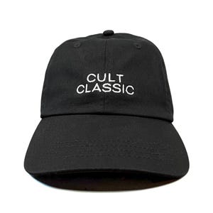 Peut inclure: Une casquette de baseball noire avec les mots CULT CLASSIC brodés en fil blanc sur le panneau avant. La casquette présente une calotte structurée, une visière incurvée avec des coutures visibles et un bouton au sommet de la structure à six panneaux.