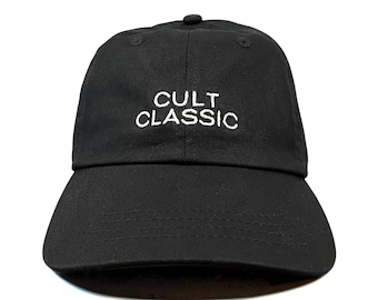 May include: A black baseball cap featuring the words CULT CLASSIC embroidered in white thread on the front panel. The cap has a structured crown, a curved brim with visible stitching, and a button at the top center of the six-panel construction.