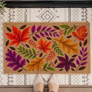 May include: A rectangular coir doormat featuring a vibrant autumn-themed print of colorful maple and oak leaves in shades of red, orange, yellow, and purple, accented with small green sprigs and red berries, layered over a white fringed rug.
