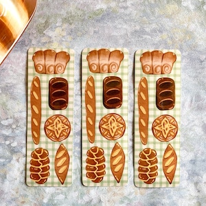 May include: Three rectangular bookmarks with illustrations of various breads, including baguettes, rolls, and loaves, on a green and white checkered background. A ceramic bread warmer is in the upper left corner.