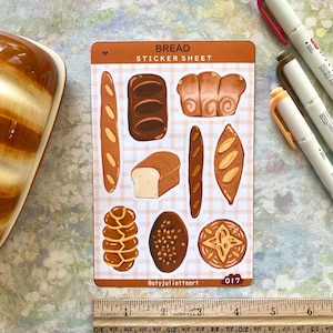 Könnte beinhalten: Ein Stickerbogen mit Illustrationen verschiedener Brotsorten, darunter Baguettes und Brotlaibe. Der Bogen hat einen braunen Hintergrund mit dem Text "BREAD STICKER SHEET" und dem Namen des Künstlers. Ein Lineal liegt darunter.