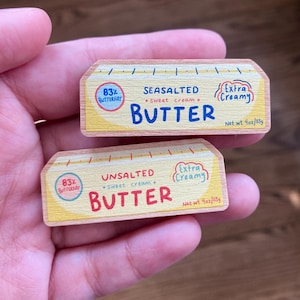 Puede incluir: Dos paquetes de madera para barras de mantequilla, uno etiquetado como "Seasalted" y el otro como "Unsalted". Ambos paquetes son amarillos con acentos rojos y azules y presentan el texto "83% Butterfat", "Sweet Cream", "Extra Creamy" y "Net wt 4oz/113g".