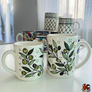 May include: White ceramic mugs with olive branch designs. The mugs feature green olives and leaves with black detailing. Other mugs with various patterns are visible in the background. The mugs have a curved handle.