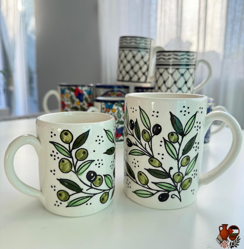 May include: White ceramic mugs with olive branch designs. The mugs feature green olives and leaves with black detailing. Other mugs with various patterns are visible in the background. The mugs have a curved handle.
