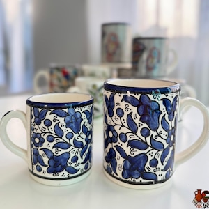 May include: Two white ceramic mugs with blue floral patterns. The mugs have a white handle and a blue rim. Additional mugs with similar designs are visible in the background.