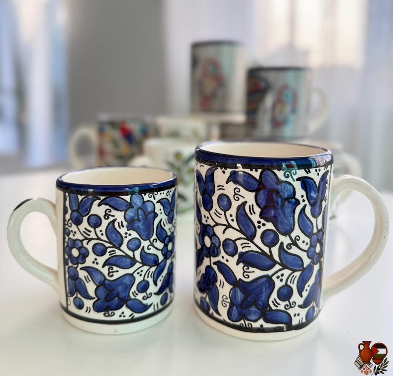 May include: Two white ceramic mugs with blue floral patterns. The mugs have a white handle and a blue rim. Additional mugs with similar designs are visible in the background.