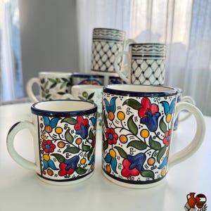 May include: A collection of ceramic mugs with colorful floral designs. The mugs feature red, blue, and yellow flowers with green leaves on a white background. Some mugs have a black and white geometric pattern.