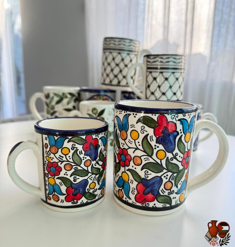 May include: A collection of ceramic mugs with colorful floral designs. The mugs feature red, blue, and yellow flowers with green leaves on a white background. Some mugs have a black and white geometric pattern.