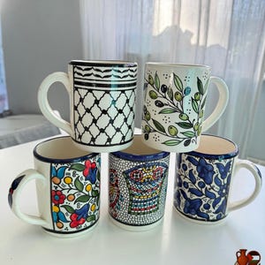 May include: Five ceramic mugs with white backgrounds and various hand-painted designs. The mugs feature patterns like black and white geometric shapes, olive branches, and floral motifs in blue, red, and yellow. Each mug has a white handle and a blue rim.