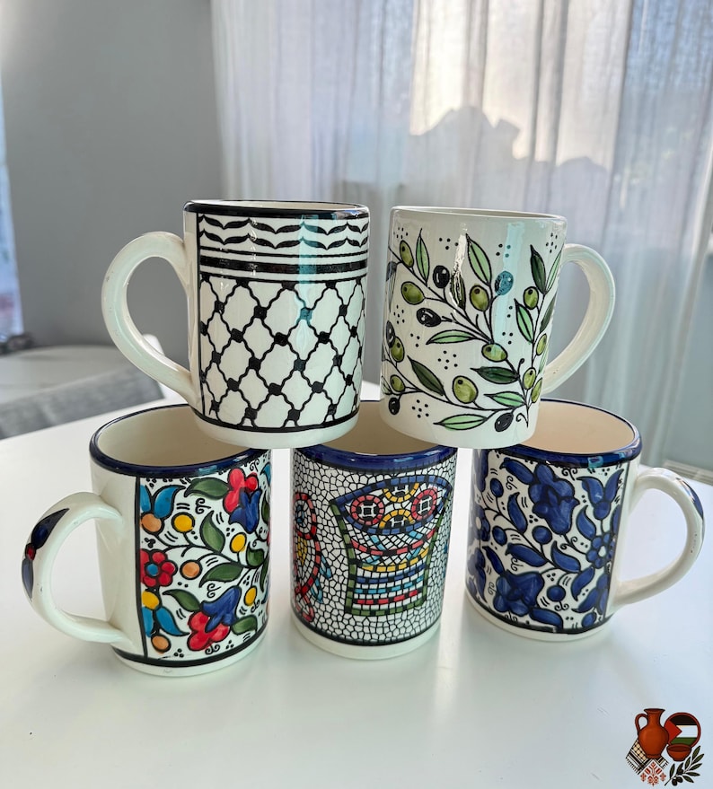 May include: Five ceramic mugs with white backgrounds and various hand-painted designs. The mugs feature patterns like black and white geometric shapes, olive branches, and floral motifs in blue, red, and yellow. Each mug has a white handle and a blue rim.