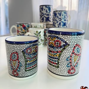 May include: A collection of ceramic mugs with various designs. The foreground features two cylindrical mugs with a mosaic-like pattern in red, yellow, blue, and green against a white and black background. Other mugs with floral and patterned designs are visible.