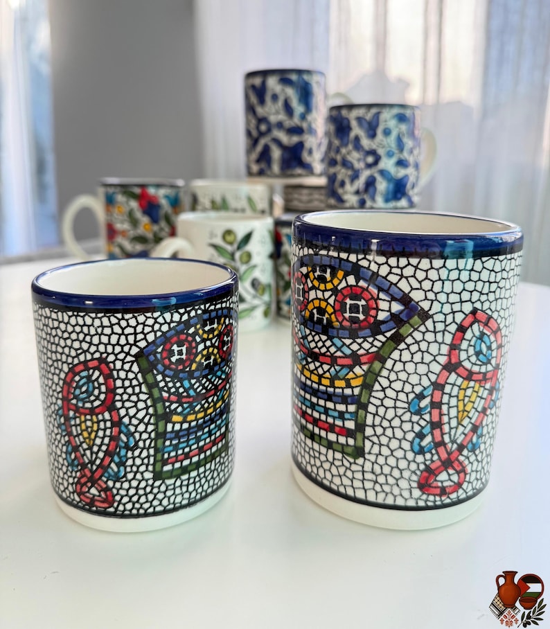 May include: A collection of ceramic mugs with various designs. The foreground features two cylindrical mugs with a mosaic-like pattern in red, yellow, blue, and green against a white and black background. Other mugs with floral and patterned designs are visible.