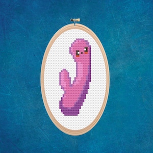 May include: A pink and purple cross-stitch embroidery of a cartoon character with a smiling face. The character is shaped like a J and has a small arm.