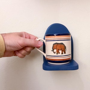 May include: A blue ceramic mug with a white elephant design and orange stripes is mounted on a blue wall-mounted shelf.