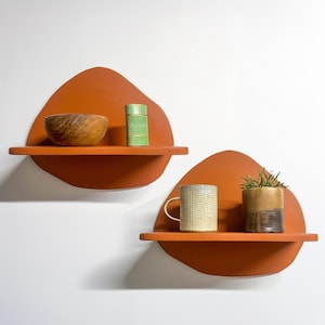 May include: Two orange wall shelves with organic shapes. One shelf holds a wooden bowl and a green container. The other shelf displays a mug and a small potted plant. The shelves are mounted on a white wall.