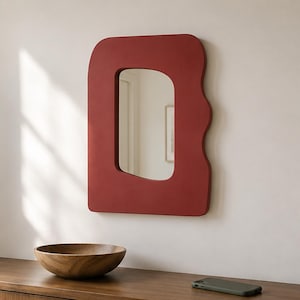 May include: A wall-mounted mirror featuring a thick, matte, deep red frame with an irregular, wavy, organic shape. The central reflective glass surface is rectangular with rounded corners, mirroring the surrounding interior space.