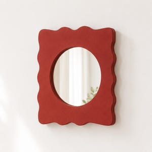 May include: A wall mirror featuring a circular glass center set within a rectangular frame that has a wavy, scalloped edge. The frame is a solid, matte terracotta red color, providing a bold contrast to the reflective circular mirror surface.