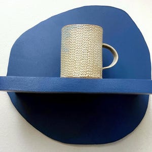 May include: A textured ceramic mug with a handle sits on a blue shelf against a blue backdrop. The mug has a beige and light blue speckled pattern. The shelf and backdrop are a solid, deep blue color. The mug is a standard size.