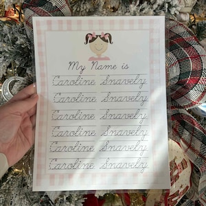 May include: A personalized name practice sheet with the text "My Name is Caroline Snavely" in cursive. The sheet has a pink gingham border and a cartoon illustration of a girl with pink bows. The sheet is held in front of a decorated Christmas tree.