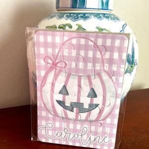May include: A small, assembled puzzle featuring a pink and white gingham pattern and a pumpkin design. The puzzle is packaged in clear plastic and has the name "Caroline" printed on it. A decorative blue and white jar is in the background.