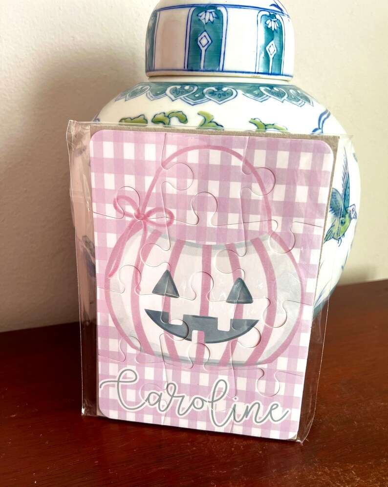 May include: A small, assembled puzzle featuring a pink and white gingham pattern and a pumpkin design. The puzzle is packaged in clear plastic and has the name "Caroline" printed on it. A decorative blue and white jar is in the background.