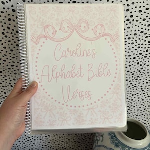 May include: A spiral-bound notebook with a pink floral design and the text "Caroline's Alphabet Bible Verses." The cover features a pink bow and floral border. The notebook is held in a person's hand.